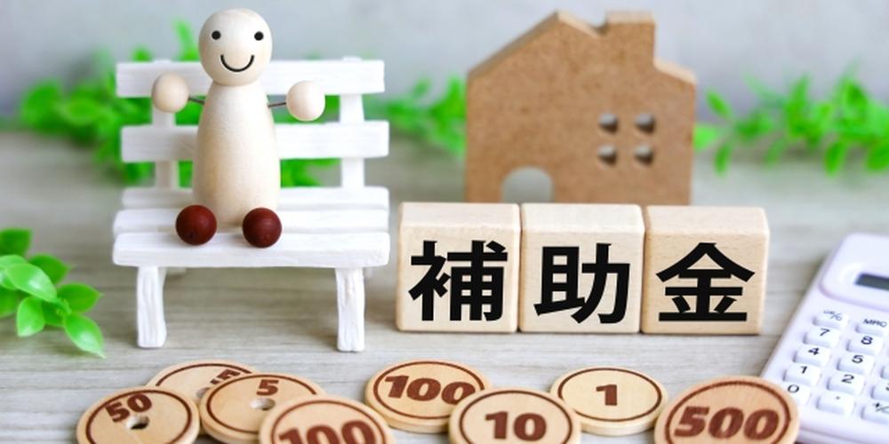 補助金をイメージした家計支援の概念画像。住宅模型と木製チップ、笑顔の人形が並ぶ。