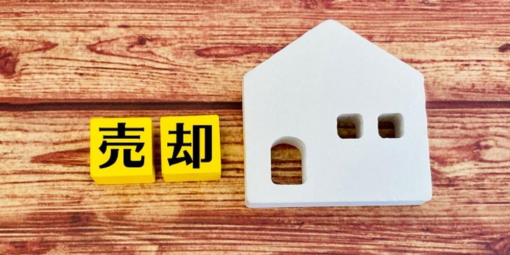 木目調の背景に置かれた「売却」の文字と白い家の模型、不動産売却をイメージした構図
